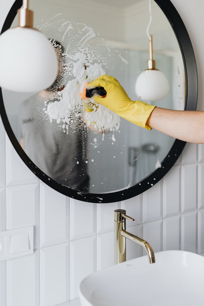 why-choose-us A person wearing a yellow glove cleans a bathroom mirror with foam spray.